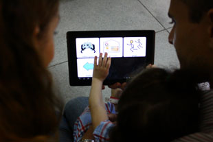 Clara manuseia o app no tablet para dizer aos pais o que quer fazer através das imagens.